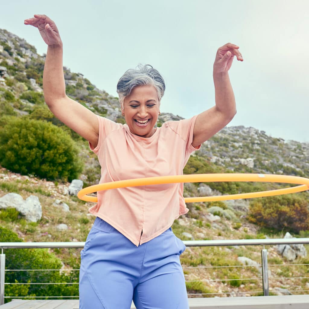 Roselani Place | Senior woman, hoop and fitness outdoor with smile, training and playful on balcony, mountains and healthy. Mature lady, exercise and happy for wellness, stomach workout and toys on rooftop in Hawaii